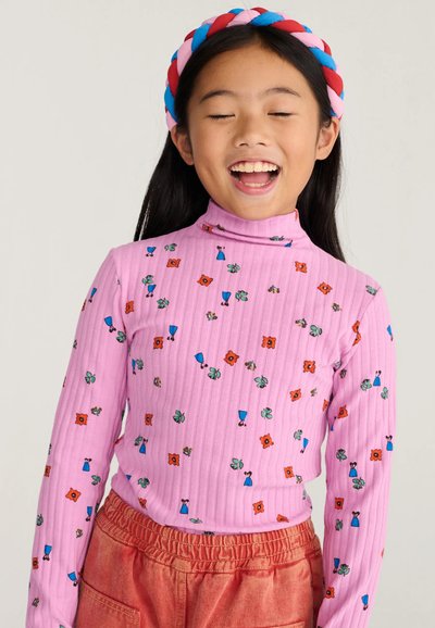 Jeune fille aux longs cheveux foncés porte une chemise rose à motifs floraux à manches longues et un pantalon rouge, souriant les yeux fermés avec un bandeau coloré.