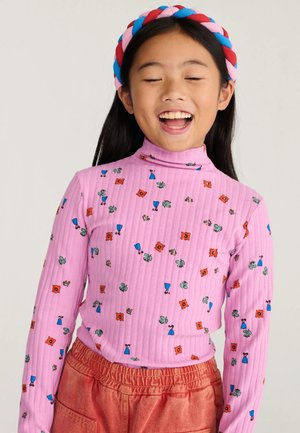 Jeune fille aux longs cheveux foncés porte une chemise rose à motifs floraux à manches longues et un pantalon rouge, souriant les yeux fermés avec un bandeau coloré.