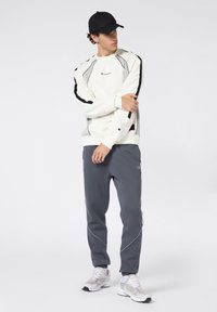 Biała bluza z czarnymi i szarymi akcentami, zestawiona z szarymi spodniami dresowymi z bocznymi lamówkami. Model nosi czapkę w kolorze czarnym i białe sneakersy.
