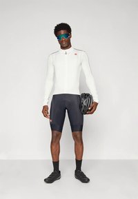 Cyklistický outfit s bielym dlhým rukávom a čiernymi šortkami. Elegantný dizajn, textúrovaná látka a červené logo. Na tvári modré slnečné okuliare.