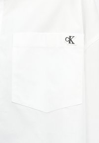 Λευκή τσέπη πουκάμισου με μικρό ασπρόμαυρο έμβλημα "cK" στην επάνω δεξιά γωνία της τσέπης.