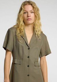 Kjole i olivengrønt med korte ermer, knappestolpe foran, klippet krage, to frontlommer og glatt stoffstruktur.