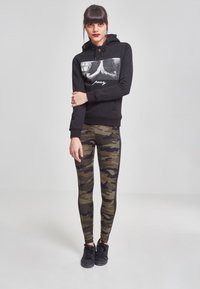Sudadera negra con estampado gráfico y texto "pray", combinada con leggings de camuflaje. Tejido texturizado y ajuste elegante. Zapatillas negras completan el conjunto.