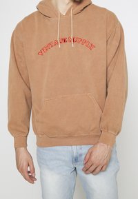 Man wearing a light brown hoodie with red "VINTAGE SUPPLY" text and light blue jeans, standing against a plain light background.