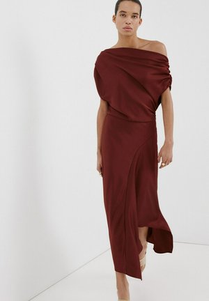 Personne portant une robe asymétrique bordeaux, épaules dénudées, avec un haut drapé et une jupe fendue, posant sur un fond blanc uni.