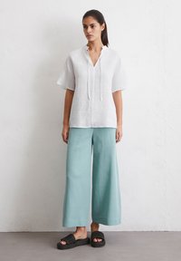 Weißes Leinenoberteil mit V-Ausschnitt und lockeren Ärmeln, kombiniert mit hellblauen weit geschnittenen Hosen. Schwarze geflochtene Sandalen runden das Outfit ab.