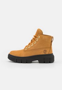 Stivaletti alla caviglia in suede tan con occhielli dorati, suola nera robusta e un'etichetta ad anello sul tallone. Presentano un logo impresso sul lato esterno.