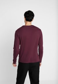 Langärmliges, bordeauxrotes T-Shirt aus leichtem Stoff, mit rundem Ausschnitt und taillierter Passform, kombiniert mit schwarzen Hosen.
