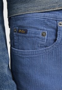 Modré kordové kalhoty s vertikálními pruhy, zdobené stříbrnými knoflíky a dekorativním štítkem "Polo" na přední kapse.