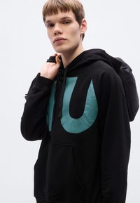 Μαύρο hoodie με μεγάλο τυρκουάζ γραφικό σχέδιο μπροστά, με τσέπη καγκουρό και ρυθμιζόμενο κορδόνι στο καπό.