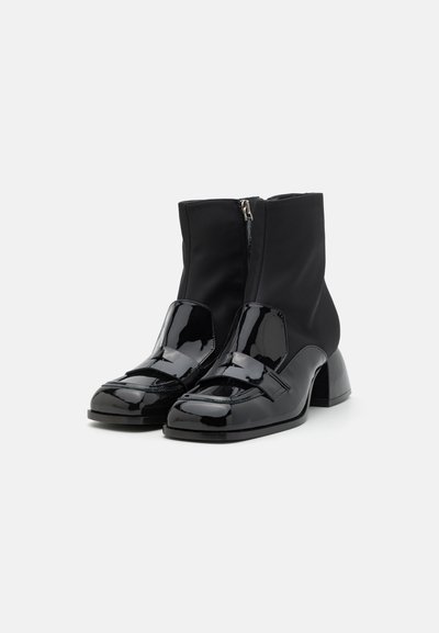 Bottines noires avec un dessus en cuir verni brillant, une tige en tissu texturé, et un talon épais avec une fermeture éclair.