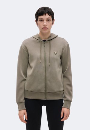 Jeune personne aux cheveux courts et foncés portant un sweat à capuche beige zippé avec des cordons et un petit logo triangulaire sur la poitrine, debout devant un fond uni.