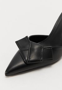 Chaussure à talons hauts en cuir noir avec un bout pointu et un détail en nœud sur le côté, présentant une texture lisse et un design épuré.