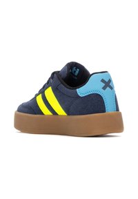 Zapatilla de ante azul marino con rayas amarillas y negras, detalles en azul claro en el talón y suela de goma marrón. Cuenta con collar y lengüeta acolchados.