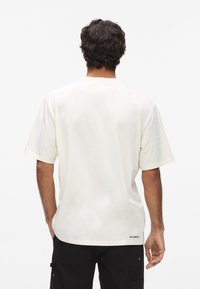 Krémové tričko s krátkymi rukávmi a okrúhlym výstrihom, vo voľnom strihu, vyrobené z bavlny, s malým čiernym logom na dolnej zadnej strane.