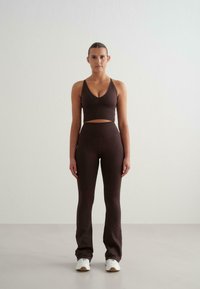 Femme debout, faisant face à l'avant, portant un débardeur court marron foncé, des leggings taille haute assortis et des baskets blanches sur un fond clair.
