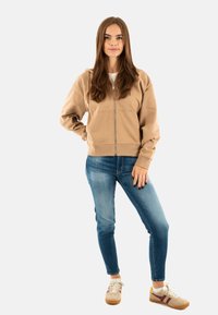 Sweat à zip marron avec manches longues, associé à un jean bleu clair et des baskets beiges avec des accents bordeaux et jaunes. Texture lisse.