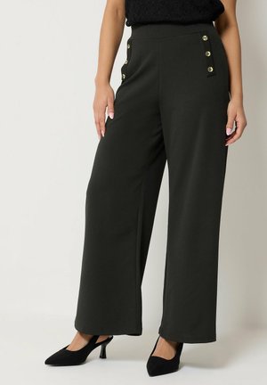 Vrouw draagt zwarte wijde hoge taillebroek met gouden knoopdetails bij de zakken, gecombineerd met zwarte slingback hakken.