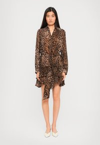 Donna in piedi che indossa un vestito lungo a maniche con stampa leopardata marrone e nera, con volant stratificati e décolleté bianchi a punta.
