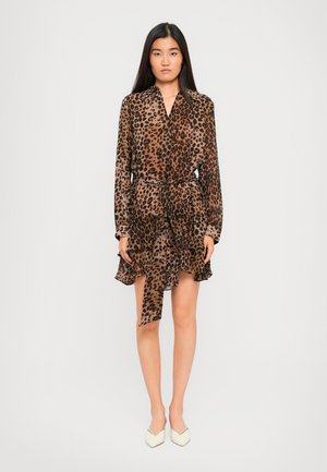 Kvinne som står iført en langermet brun og svart leopardmønstret kjole med lagdelte volanger og hvite spisse hæler.