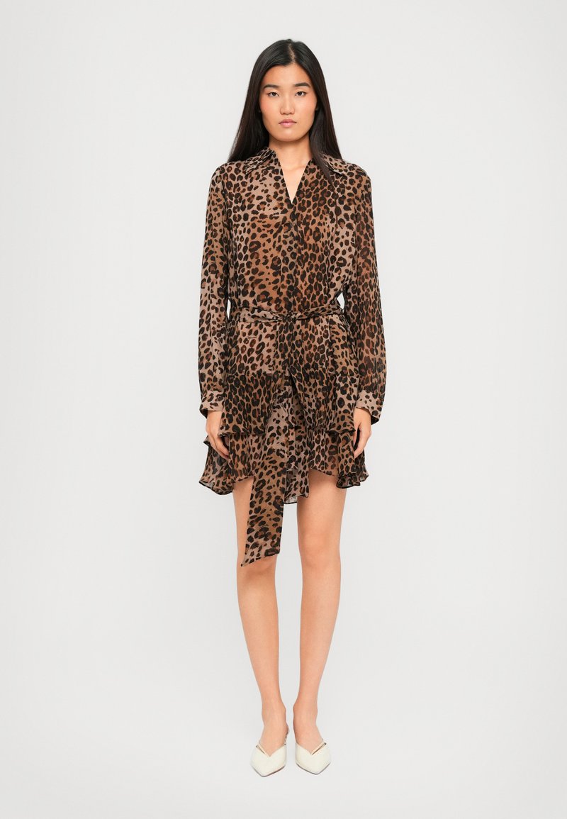Donna in piedi che indossa un vestito lungo a maniche con stampa leopardata marrone e nera, con volant stratificati e décolleté bianchi a punta.