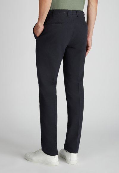 Man in zwarte slim-fit trousers met achterzakken en witte sneakers, staand met één hand in zijn zak tegen een effen achtergrond.