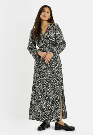 Mujer con un vestido maxi de manga larga blanco y negro con manchas, abertura frontal y mocasines negros, posando frente a un fondo blanco.