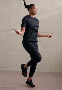 Schwarzes Sport-T-Shirt und Leggings, getragen von einer Person, die Seilspringen macht. Schwarze Sneaker mit rosa Akzenten, glatte Texturen, Innenraum.