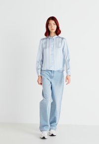 Chemise bleu satin clair à boutons avec des rayures verticales, associée à un jean baggy bleu clair et des baskets blanches. Texture lisse et coupe décontractée.