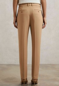 Pantaloni beige su misura con vestibilità dritta, dotati di due tasche posteriori con bottoni e una texture liscia. Indossati con scarpe con tacco alto.