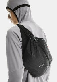 Henkilö, joka käyttää vaaleanharmaata hupparia ja mustaa olkataskua, jossa on vetoketju ja logo takana, katsoo vasemmalle neutraalissa taustassa.