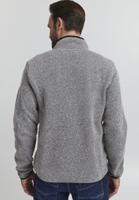 Pulover gri din fleece cu guler înalt și finisaj texturat. Spatele are mâneci lungi și croială dreaptă, asociat cu blugi de culoare bleu închis.