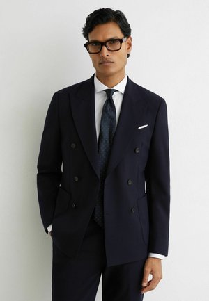 Mand iført sort dobbeltradet jakkesæt, hvid skjorte, mørkeblå slips med små hvide prikker og sorte briller, stående foran en hvid væg.