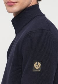 Pull à fermeture éclair bleu marine avec col haut, texture côtelée, et un patch logo Belstaff noir et or distinctif sur le haut du bras.
