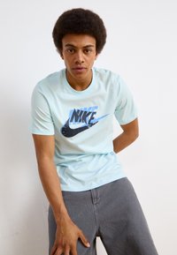 Světle modré bavlněné tričko s výrazným černým a modrým logem Nike na přední straně. Krátké rukávy, kulatý výstřih, volný střih.