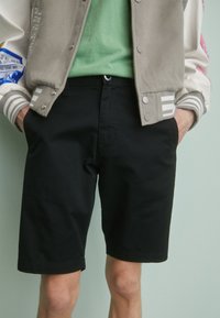 Shorts en coton noirs à coupe classique avec poches avant, associés à un T-shirt vert clair et une veste de type varsity beige avec des accents colorés.