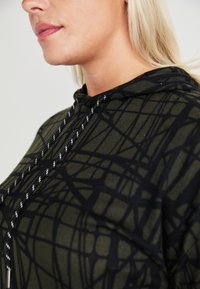 Svetlana s kapuco v temno zeleni barvi z črnim abstraktnim vzorcem. Ima kapuco s vrvico in teksturirano tkanino. Oblika je sproščena.