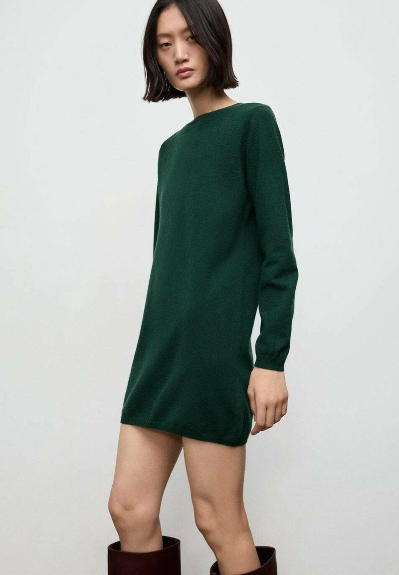 Abito verde a maglia con maniche lunghe, taglio dritto e scollatura rotonda. Tessuto liscio, lunghezza fino a metà coscia, abbinato a stivali marroni al ginocchio.