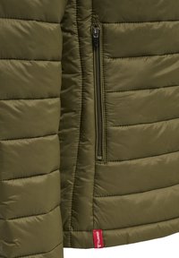 Close-up van olijfgroene doorgestikte jas met verticale ritssluiting en klein rood merkembleem onderaan de zoom.
