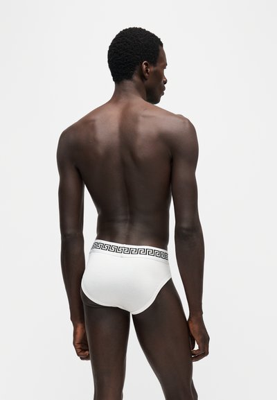 Homme de dos à la caméra, portant un slip blanc avec une ceinture noire à motif grec, sur un fond uni et clair.