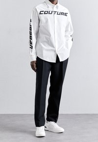 Weißes Hemd mit Knöpfen, vorne und an den Ärmeln mit schwarzem "COUTURE"-Aufdruck, kombiniert mit schwarzer Hose und weißen Sneakers. Glatte Stoffstruktur.