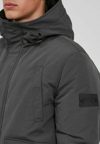 Gray padded jacket with a high collar, drawstring hood, side pockets, and a black patch logo on the left sleeve. Textured and durable material.