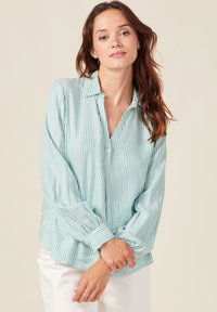 Blusa a righe verde chiaro con colletto, maniche lunghe e patta con bottoni. Tessuto morbido con una vestibilità comoda, abbinata a pantaloni bianchi.