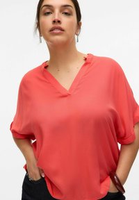 Blusa de color coral con escote en V, mangas cortas y corte holgado. Confeccionada en tela liviana, presenta un suave drapeado y detalles de mangas enrolladas.