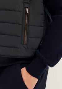 Schwarze Steppweste mit einer zipfeligen Brusttasche, braunen Reißverschlussakzenten und einem darunter teilweise sichtbaren strukturierten marineblauen Pullover.
