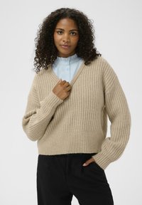 Beżowy sweter dzianinowy z dekoltem w kształcie V, struktura ribbed, opadające ramiona i szerokie rękawy. W zestawieniu z jasnoniebieską koszulą z kołnierzykiem.