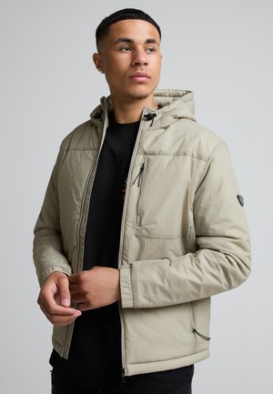Ung mand iført en beige hættejakke over en sort skjorte, der kigger til siden med hænderne nær jakke lynlåsen.