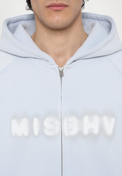 Lys blå zip-up hettegenser med en myk tekstur, med et fremtredende hvitt "MISBHV"-logo i midten og en sølvglinsende glidelås.