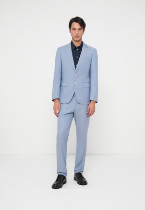 Costume ajusté bleu clair avec une veste à un bouton et un pantalon slim, associé à une chemise noire à boutons et des chaussures noires.