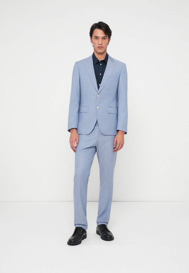 Šviesiai mėlynas siuvinėtas kostiumas su viena saga, siaura kelnėmis, derinamas su tamsiai mėlyna marškiniais ir juodais batais.